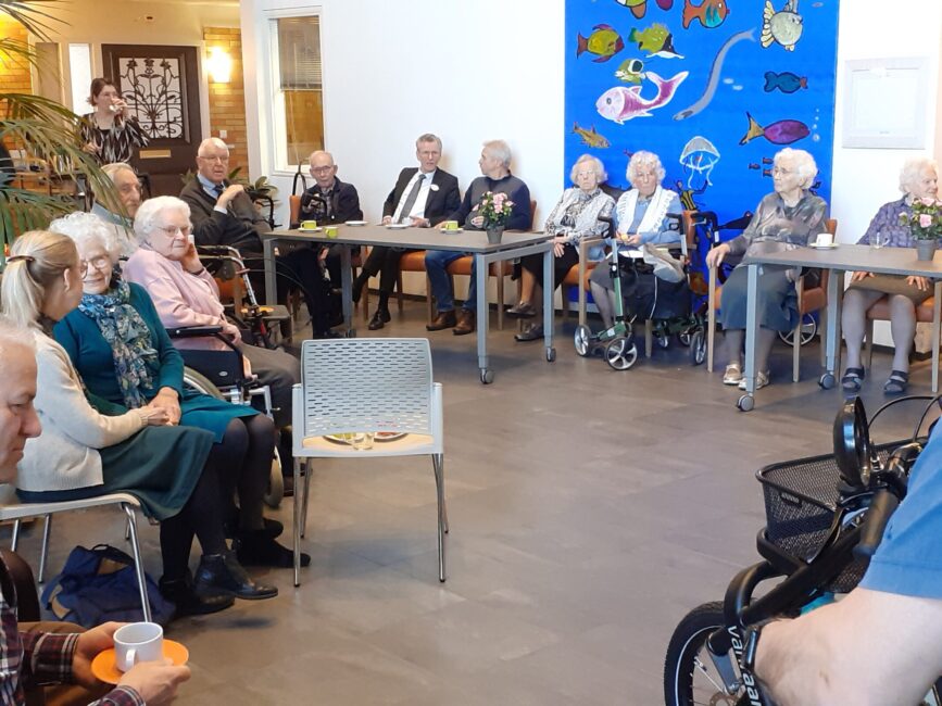 Groep ouderen zit in een gemeenschappelijke ruimte, luisterend naar een spreker. Achter hen hangt een kleurrijk viskunstwerk.
