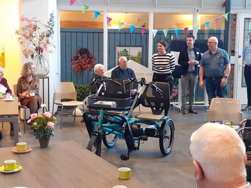 Groep mensen in een zaal met een blauwe duofiets in het midden. Er hangen kleurrijke vlaggetjes aan het plafond.