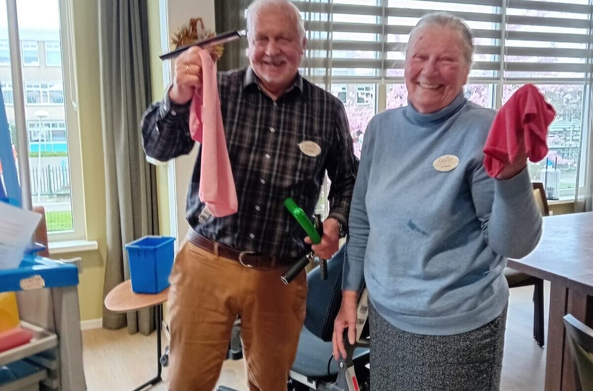 Twee lachende mensen in een kamer, houden schoonmaakdoeken en gereedschap vast. Op de achtergrond staat een rolstoel.