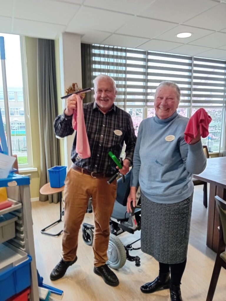 Twee lachende mensen in een kamer, houden schoonmaakdoeken en gereedschap vast. Op de achtergrond staat een rolstoel.
