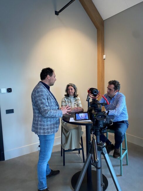 Drie mensen in gesprek tijdens een mediatraining. Een man staat, terwijl de anderen aan een tafel zitten met opnameapparatuur.
