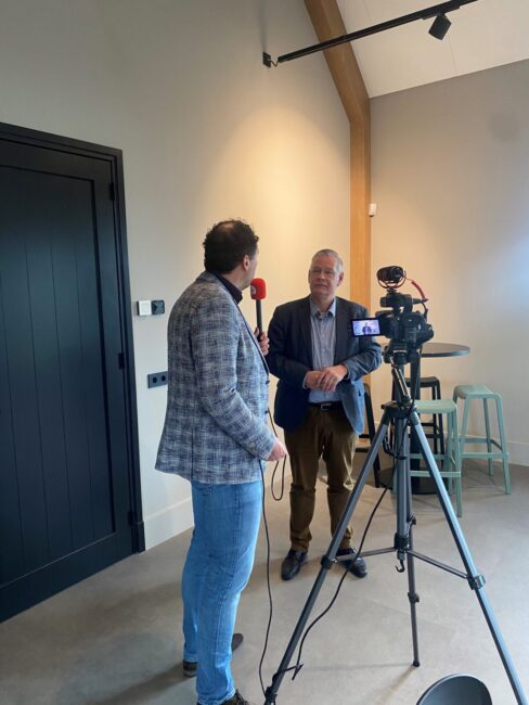 Twee mannen in gesprek voor een camera tijdens een mediatraining in een moderne ruimte met een hoge tafel en krukken.