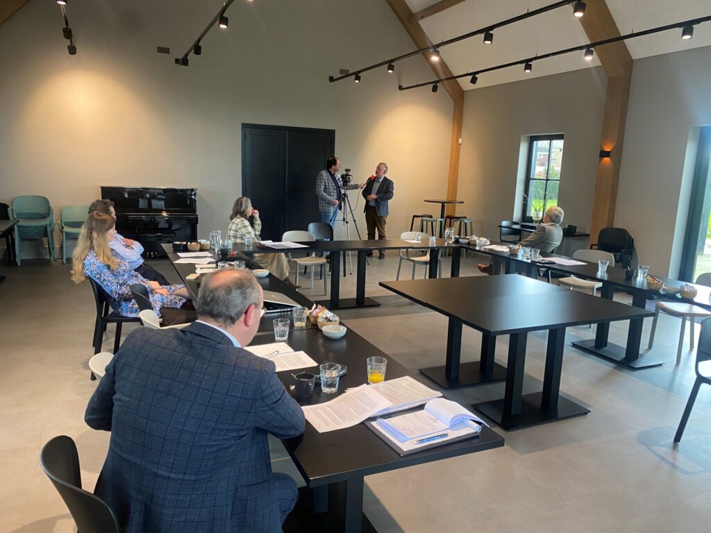 Een groep mensen zit in een vergaderruimte met tafels en stoelen, terwijl een presentatie wordt gegeven aan de voorkant van de zaal.