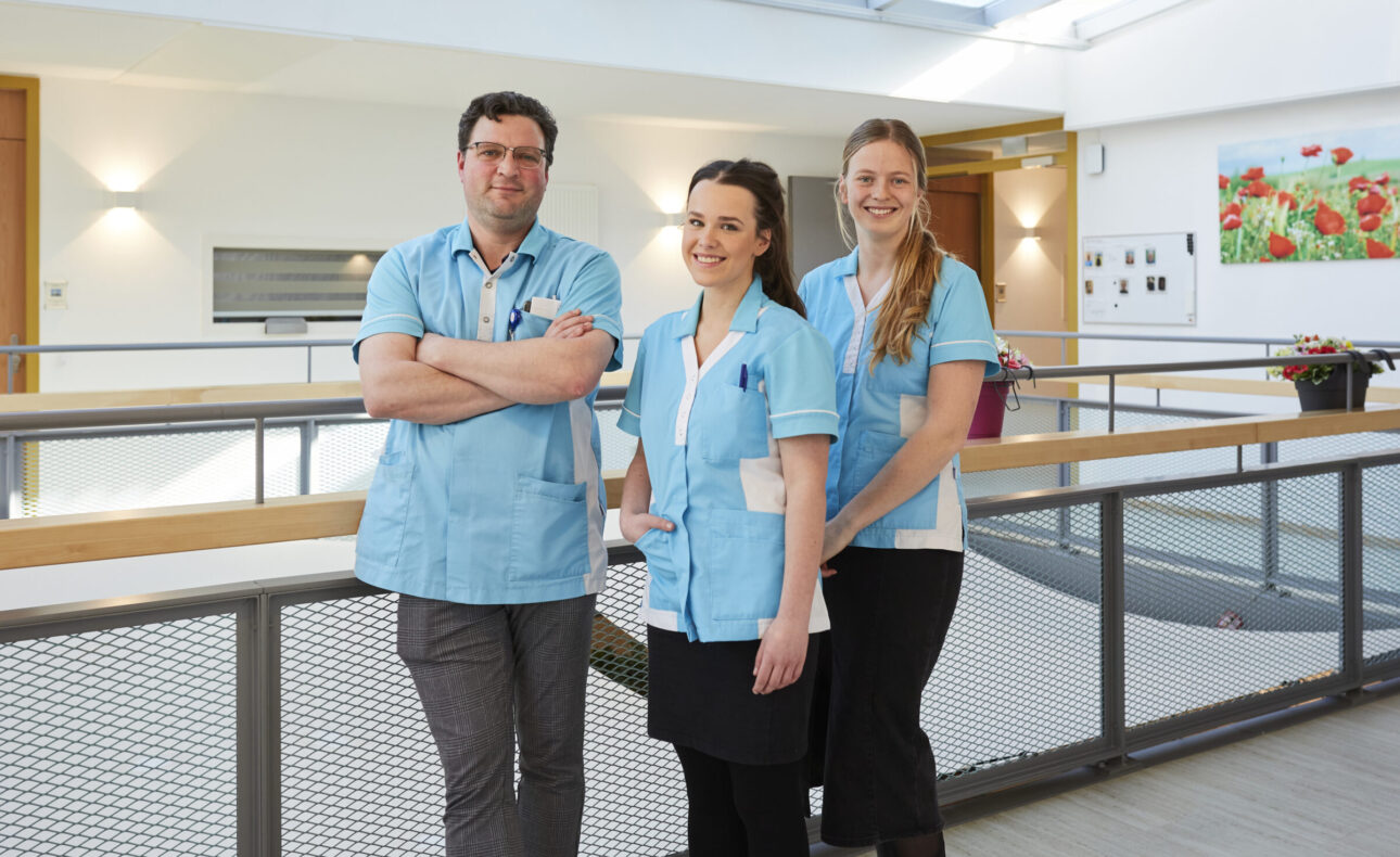 Drie zorgmedewerkers in blauwe uniformen staan glimlachend in een lichte gang van een zorginstelling, met kunst aan de muur.