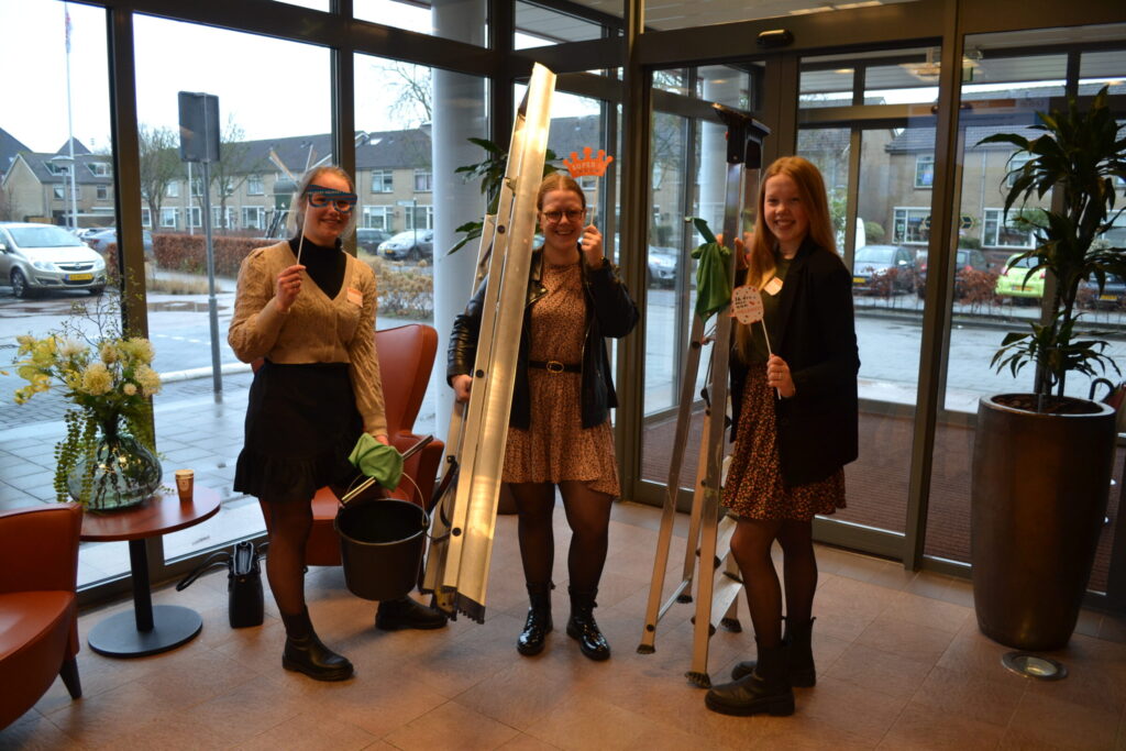 Drie vrouwen staan in een hal met glazen deuren, houden planten en ladders vast, en lachen naar de camera. Buiten is het bewolkt.
