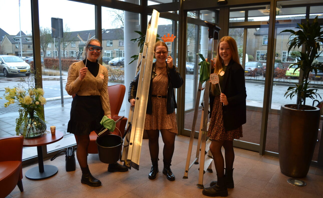 Drie vrouwen staan in een hal met glazen deuren, houden planten en ladders vast, en lachen naar de camera. Buiten is het bewolkt.