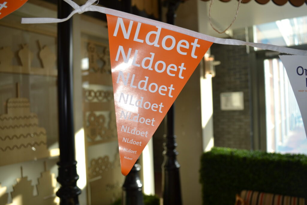 Oranje vlaggetjes met de tekst "NLdoet" hangen aan een lijn. Ze zijn onderdeel van een decoratie in een zonnige ruimte.
