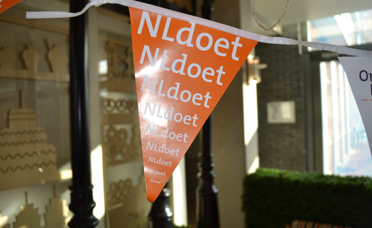 Oranje vlaggetjes met de tekst "NLdoet" hangen aan een lijn. Ze zijn onderdeel van een decoratie in een zonnige ruimte.