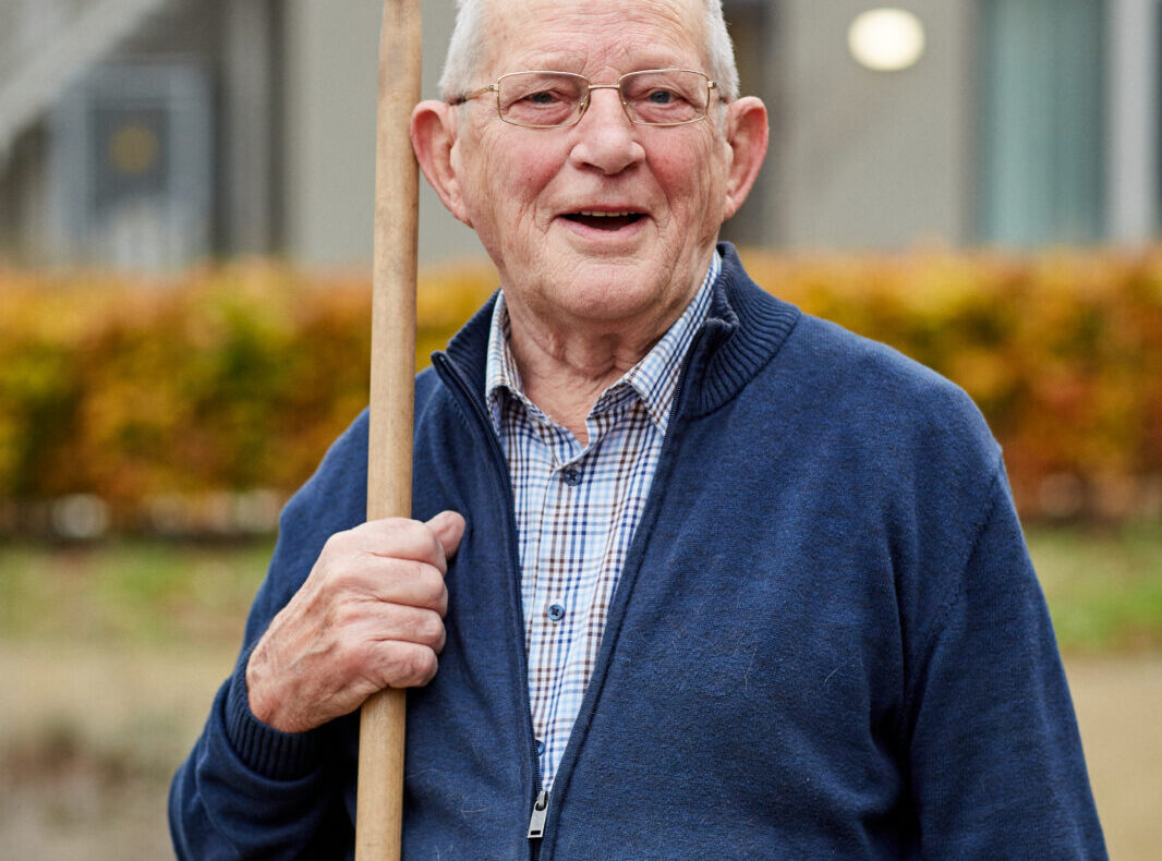 Oudere man met bril en wandelstok, glimlachend buiten voor een gebouw, gekleed in een blauwe trui en geruite overhemd.