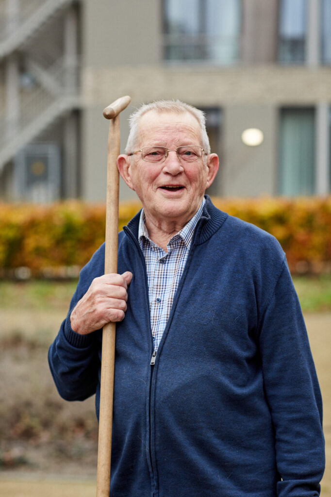 Oudere man met bril en wandelstok, glimlachend buiten voor een gebouw, gekleed in een blauwe trui en geruite overhemd.