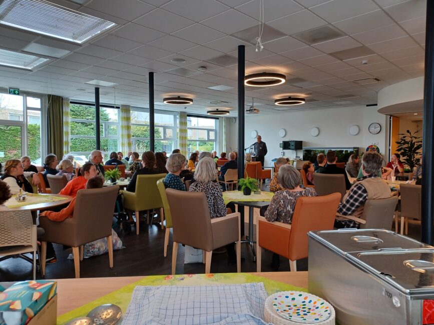 Groep mensen zit in een lichte ruimte met tafels en stoelen, luisterend naar een spreker. Er zijn grote ramen en planten.