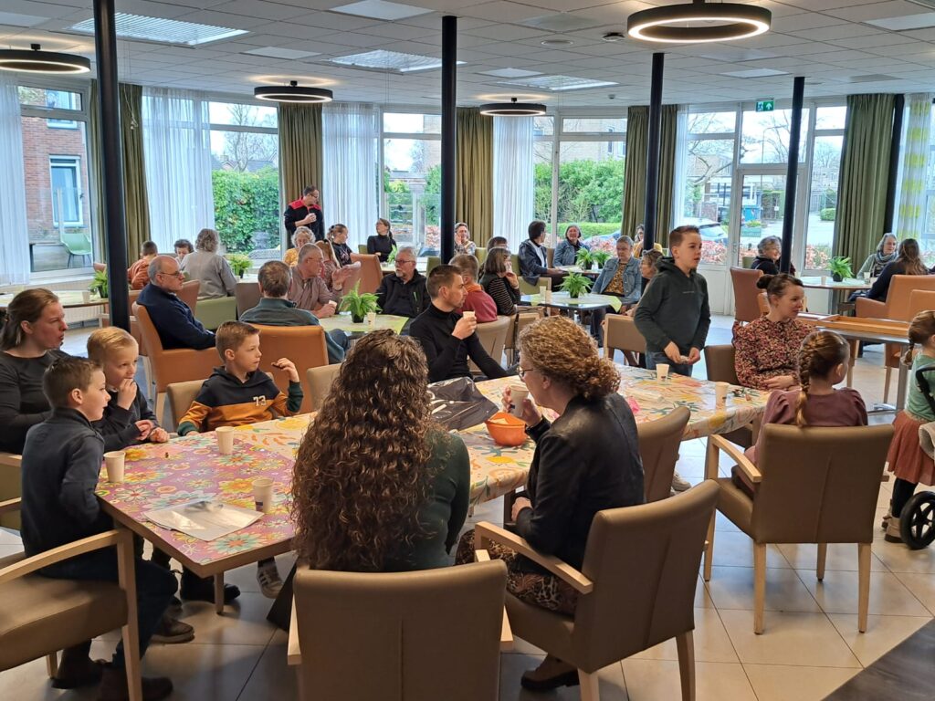 Groep mensen van verschillende leeftijden zit aan tafels in een lichte ruimte met grote ramen, bezig met sociale activiteiten.