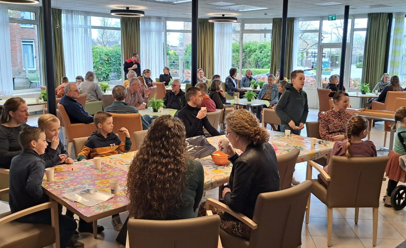 Groep mensen van verschillende leeftijden zit aan tafels in een lichte ruimte met grote ramen, bezig met sociale activiteiten.