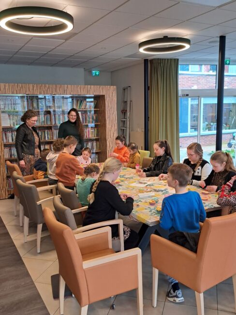 Groep kinderen knutselt aan tafel in een bibliotheek, begeleid door twee volwassenen. Boekenplanken op de achtergrond.