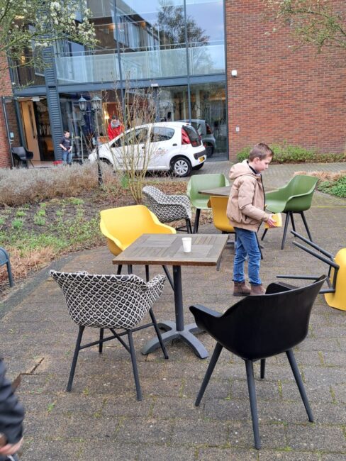 Een kind speelt buiten bij een tafel met stoelen op een terras. Op de achtergrond is een gebouw met grote ramen zichtbaar.