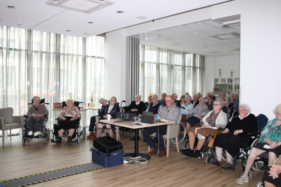 Een groep ouderen zit in een lichte ruimte, aandachtig kijkend naar een presentatie. Sommigen zitten in rolstoelen.