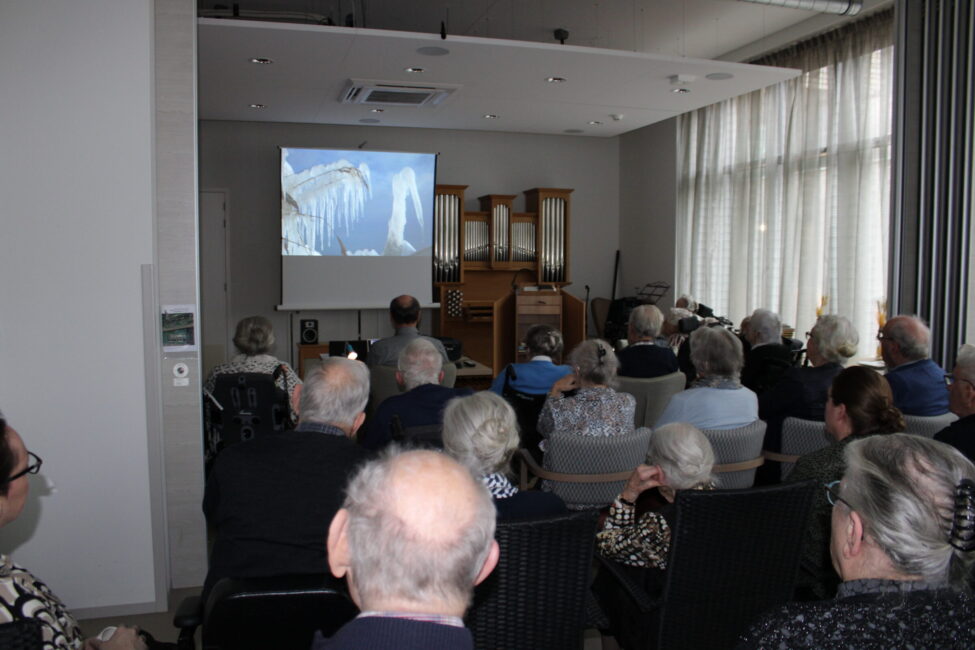 Een groep mensen kijkt naar een projectiescherm in een zaal. Op het scherm is een afbeelding van een waterval te zien.