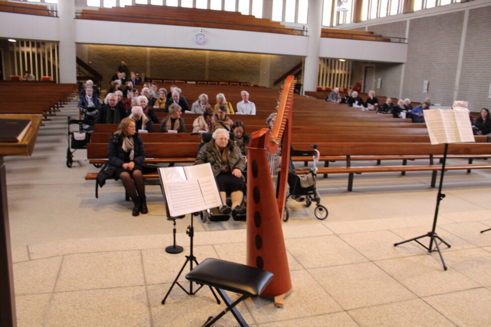 Een harp en muziekstandaards staan klaar in een kerkzaal met houten banken. Enkele mensen zitten in afwachting van een optreden.