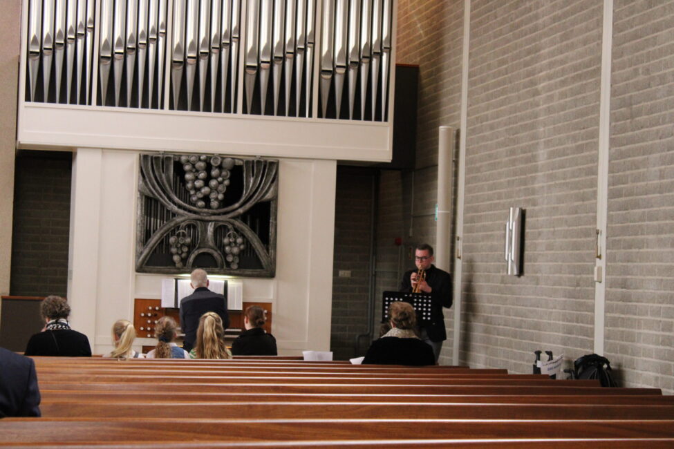 Een groep mensen zit in kerkbanken, terwijl een man vooraan bij een orgel staat. De kerk heeft een moderne uitstraling.