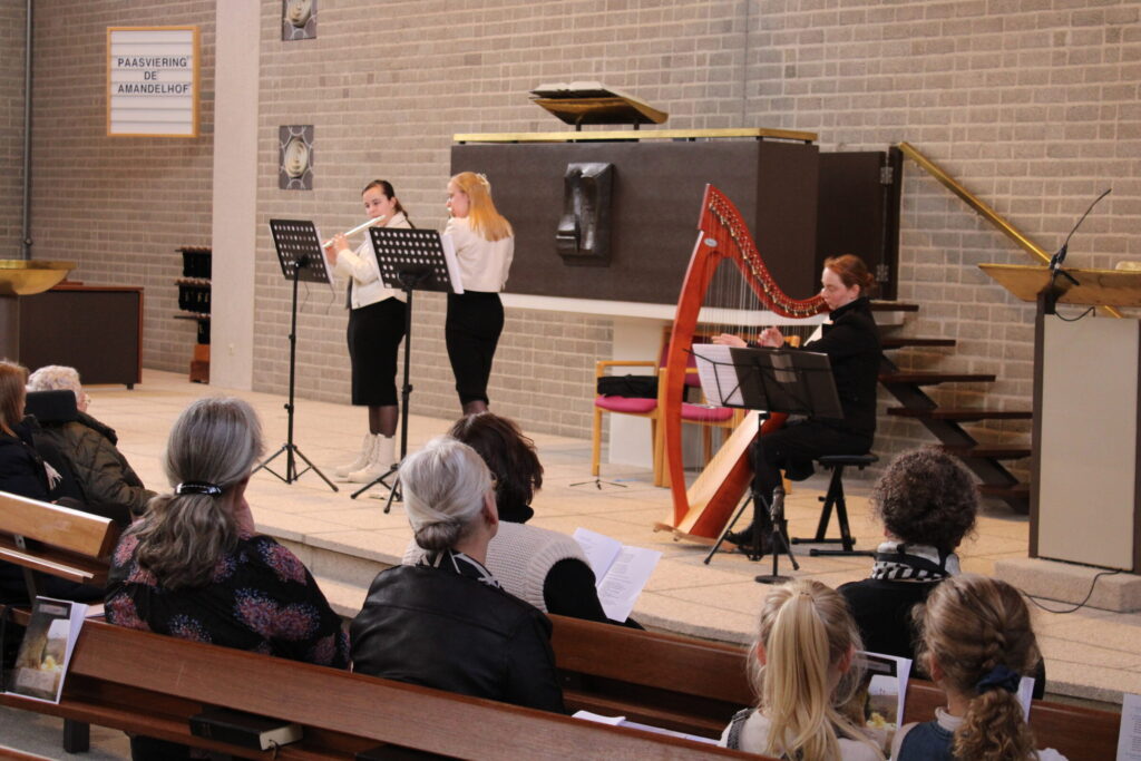Muzikanten spelen fluit en harp voor een zittend publiek in een zaal met houten banken en een groot orgel op de achtergrond.