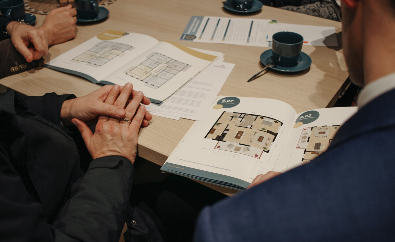 Twee mensen bekijken plattegronden in een boek aan een tafel met koffiekopjes en papieren. Ze bespreken de inhoud samen.