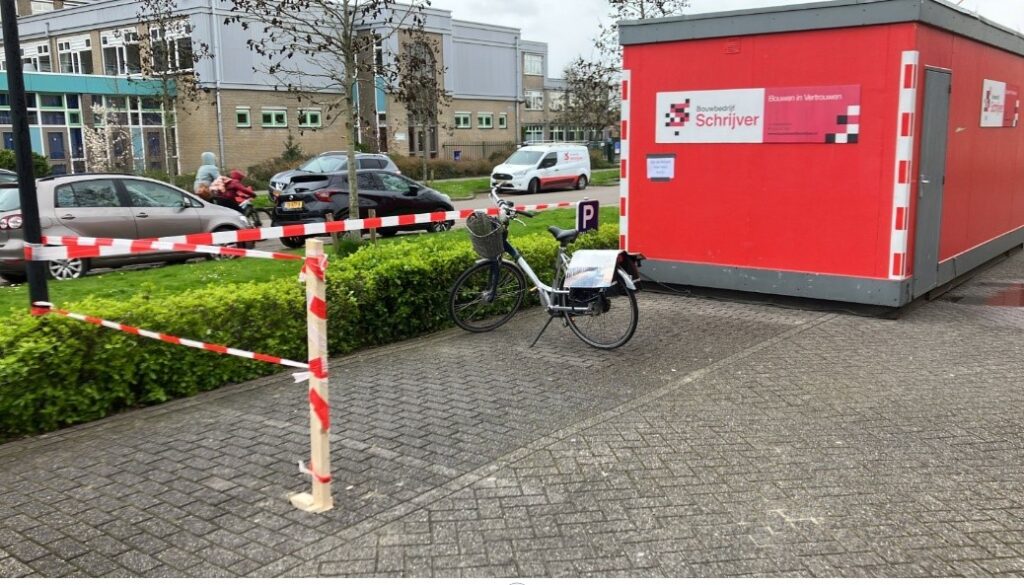 Fiets geparkeerd naast een rood gebouw met een bord van een zorginstelling. Er zijn auto's en een parkeerplaats zichtbaar.