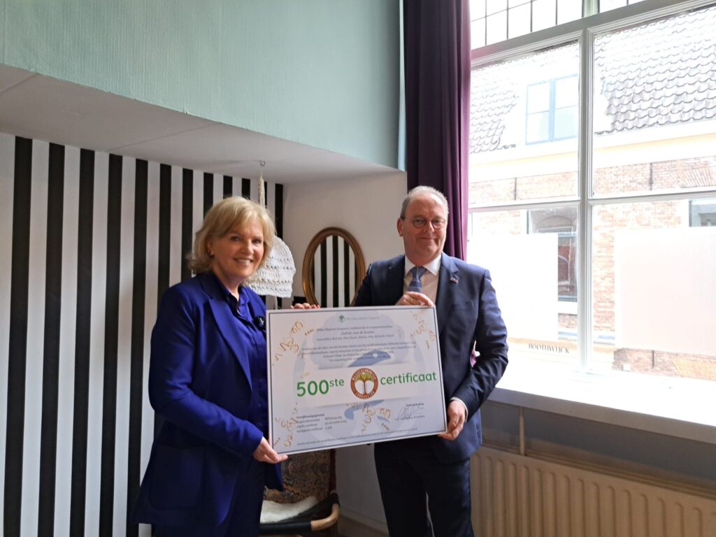 Twee mensen houden een groot certificaat vast in een kamer met een raam en gestreepte muur. Ze lijken blij en trots.