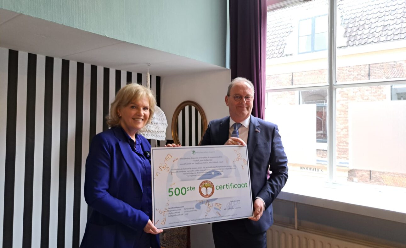Twee mensen houden een groot certificaat vast in een kamer met een raam en gestreepte muur. Ze lijken blij en trots.