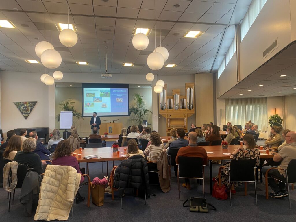 Een groep mensen zit in een zaal en luistert naar een presentatie. Er hangen ronde lampen aan het plafond en een scherm is zichtbaar.