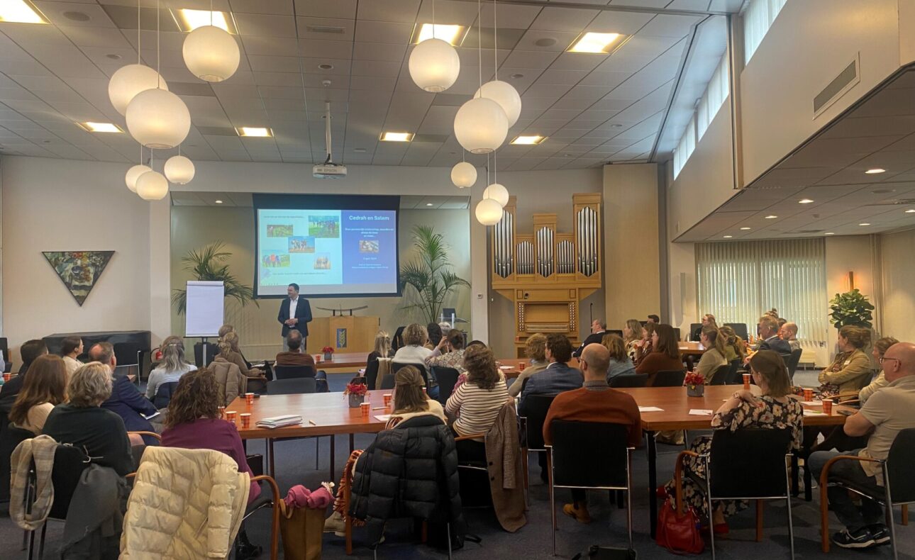 Een groep mensen zit in een zaal en luistert naar een presentatie. Er hangen ronde lampen aan het plafond en een scherm is zichtbaar.