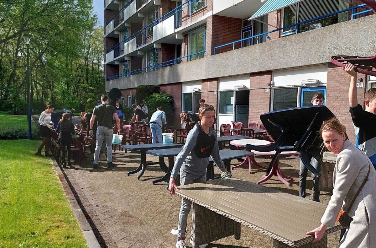Groep scholieren verplaatst tafels buiten bij een flatgebouw op een zonnige dag, omringd door groen en blauwe lucht.