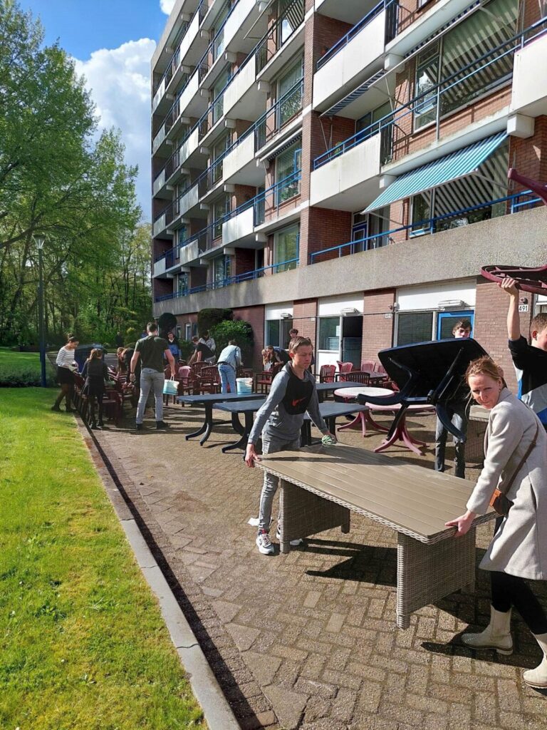 Groep scholieren verplaatst tafels buiten bij een flatgebouw op een zonnige dag, omringd door groen en blauwe lucht.