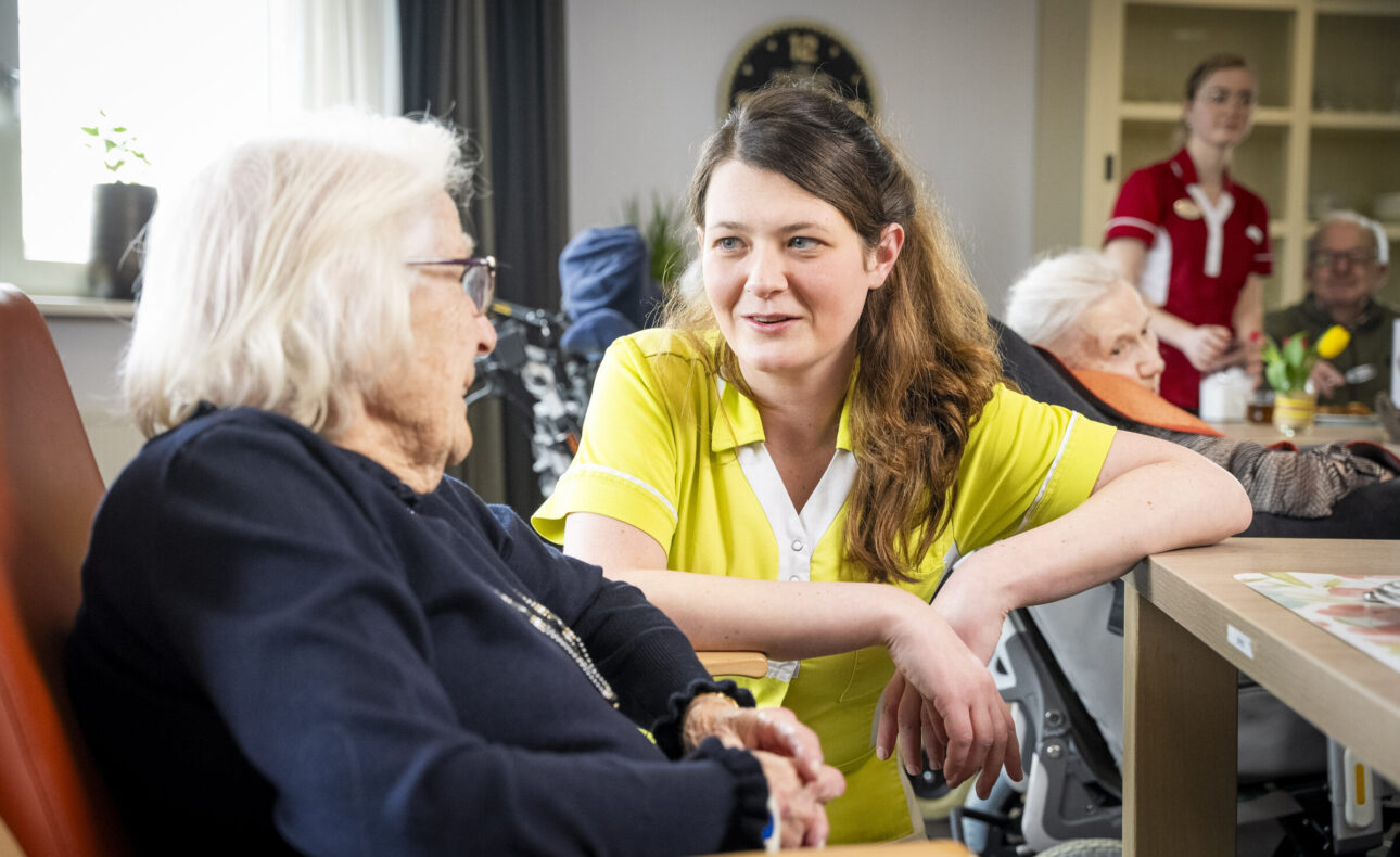 Twee vrouwen in gesprek in een zorginstelling. Een zorgmedewerker in gele kleding praat met een oudere vrouw in een stoel.