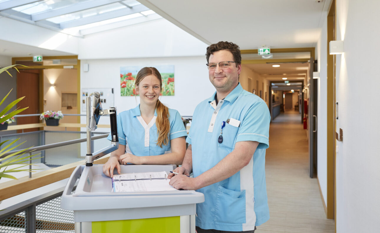 Twee zorgmedewerkers in blauwe uniformen staan naast een medische kar in een lichte gang van een zorginstelling.