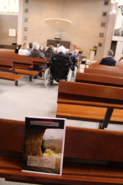 Een kerkinterieur met houten banken. Op de voorgrond ligt een boekje op een bank. Mensen zitten verderop in de kerk.