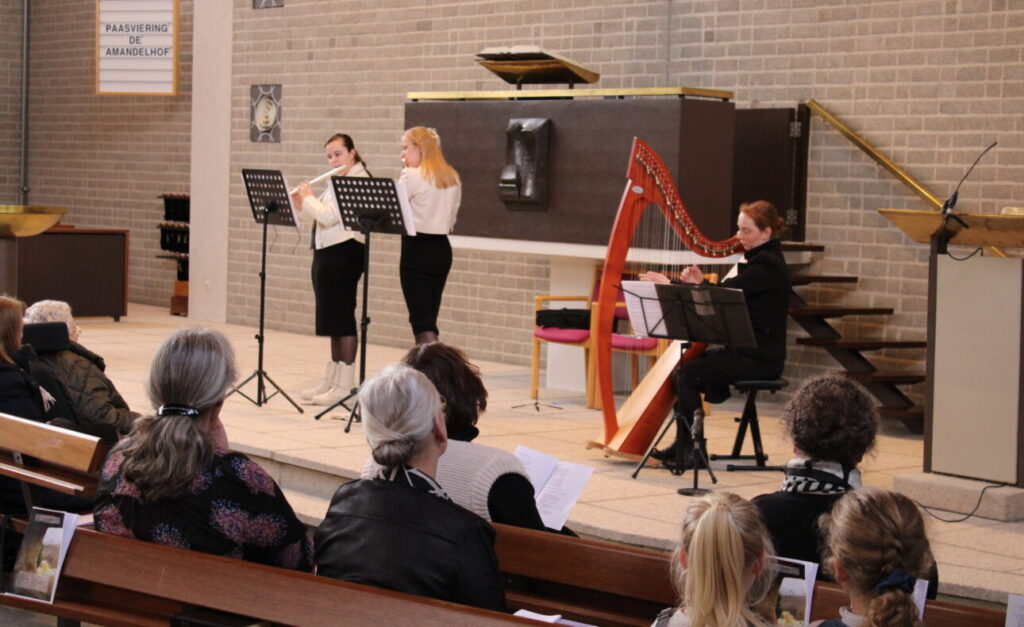 Muzikanten spelen fluit en harp voor een zittend publiek in een kerk. Mensen luisteren aandachtig naar het optreden.