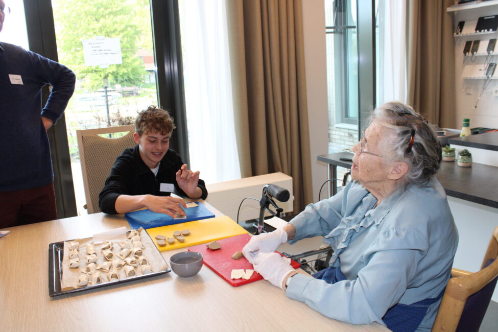 Twee mensen zitten aan een tafel en bereiden koekjes voor. Een oudere vrouw draagt een schort en handschoenen, een jongere persoon lacht.