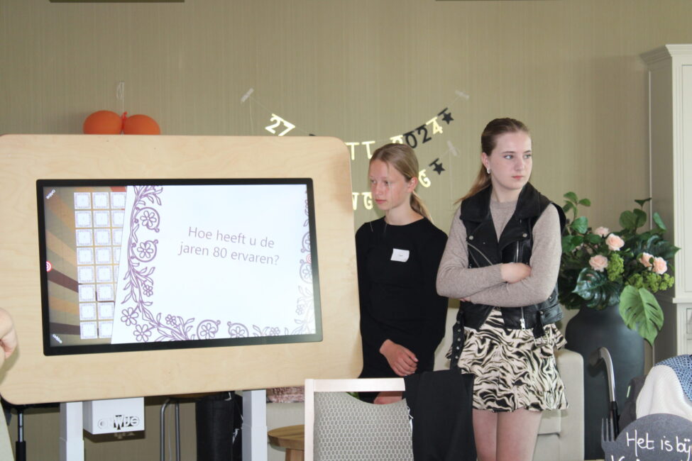 Twee jonge vrouwen staan naast een scherm met een presentatie in een kamer. Er zijn decoraties en planten op de achtergrond.