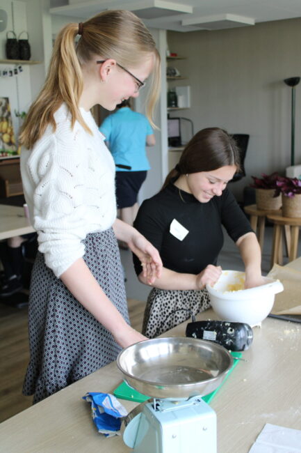 Twee jonge vrouwen bakken samen in een keuken. De ene vrouw mengt ingrediënten in een kom, de ander kijkt toe en lacht.