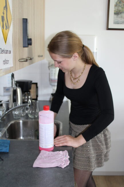 Vrouw in zwarte top en geruite rok maakt het aanrecht schoon met een roze doek en schoonmaakmiddel in een keuken.