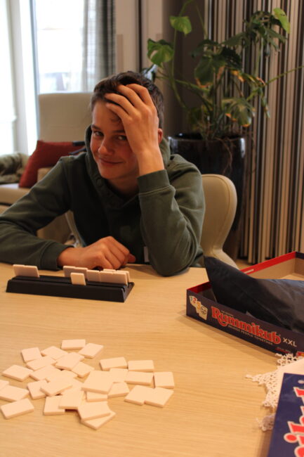 Een persoon zit aan een tafel met een bordspel en stenen. Ze glimlachen en leunen met hun hand tegen hun hoofd.