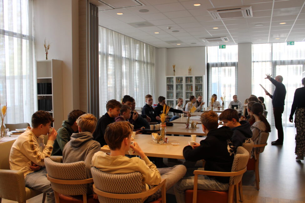 Groep jongeren zit aan tafels in een lichte ruimte met grote ramen, terwijl een persoon vooraan iets presenteert of uitlegt.