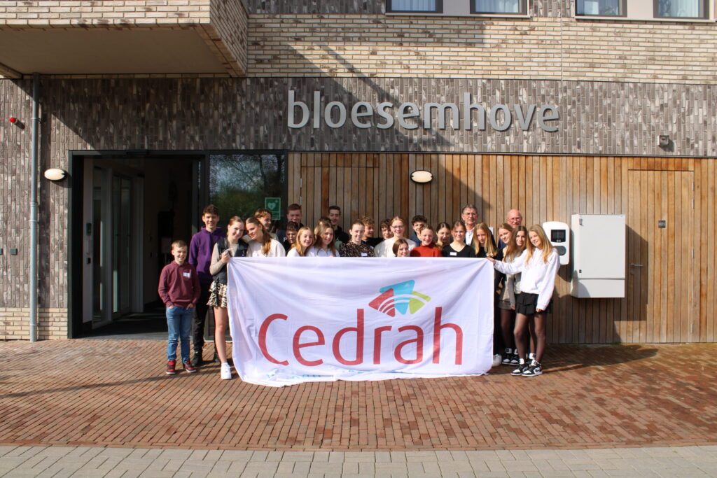 Groep mensen poseert voor een gebouw met een bord "bloesemhof", terwijl ze een groot spandoek met het logo van Cedrah vasthouden.