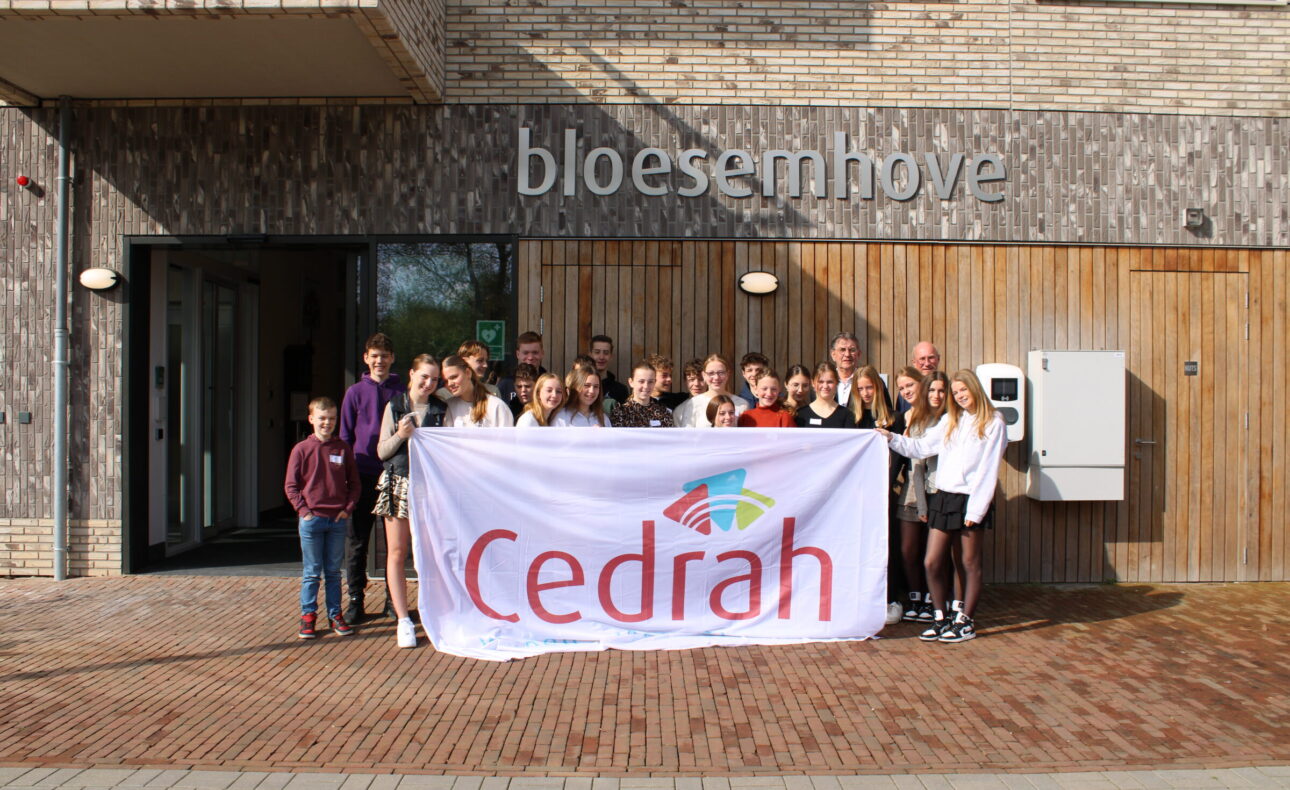 Groep mensen poseert voor een gebouw met een bord "bloesemhof", terwijl ze een groot spandoek met het logo van Cedrah vasthouden.