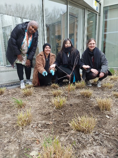 Vier mensen poseren buiten voor een gebouw, gehurkt bij een tuin met jonge planten. Ze dragen warme kleding en glimlachen.