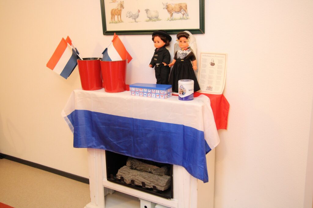 Tafel met Nederlandse vlag, poppen in traditionele klederdracht, en decoratieve items, waaronder vlaggetjes en een certificaat.