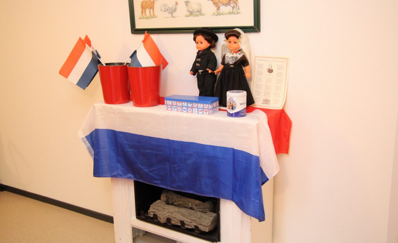 Tafel met Nederlandse vlag, poppen in traditionele klederdracht, en decoratieve items, waaronder vlaggetjes en een certificaat.