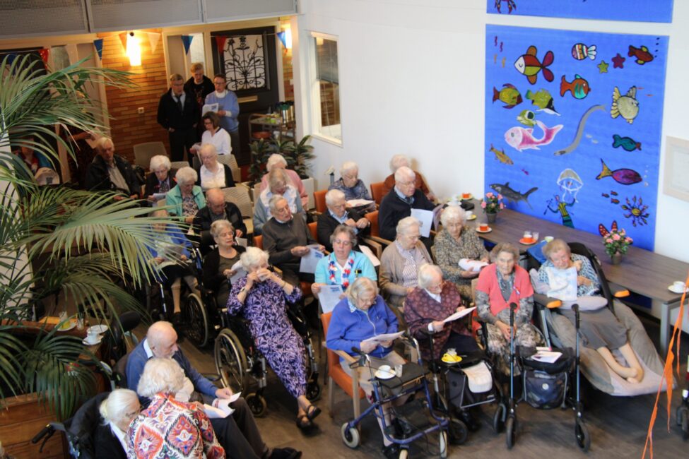 Groep ouderen in een gemeenschappelijke ruimte, zittend in rolstoelen en stoelen, met kleurrijke muurschildering op de achtergrond.
