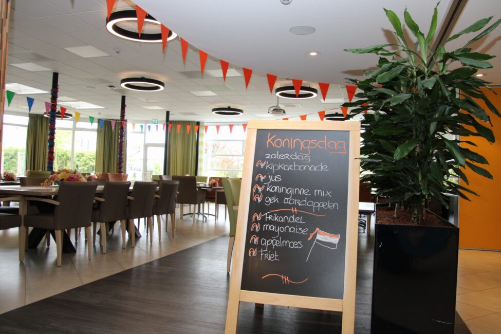 Een feestelijk versierde eetzaal met oranje vlaggetjes en een krijtbord met het programma voor Koningsdag.