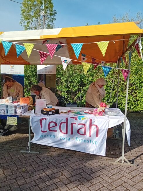 Kraam met kleurrijke vlaggetjes en Cedrah-banner. Mensen staan achter de tafel met folders en producten op een zonnige dag.